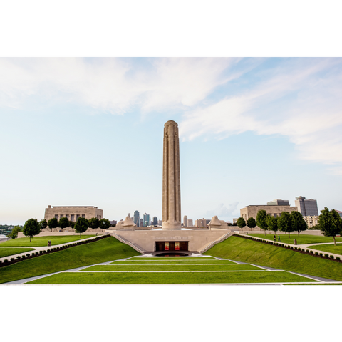 The image shows the Liberty Memorial, part of the National WWI Museum & Memorial, a tall stone tower surrounded by landscaped gardens and two large buildings, with a city skyline in the background.