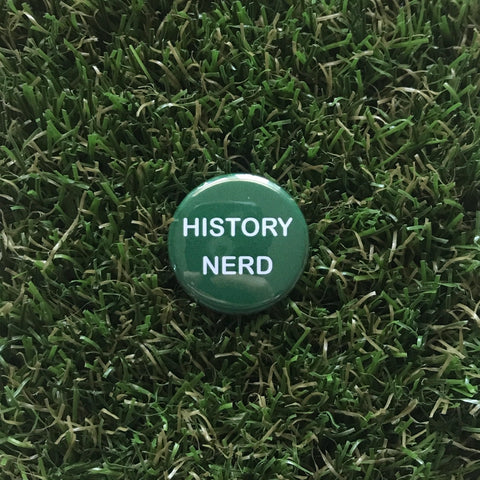 A hand-pressed green button with History Nerd elegantly printed on it rests atop lush green artificial grass.