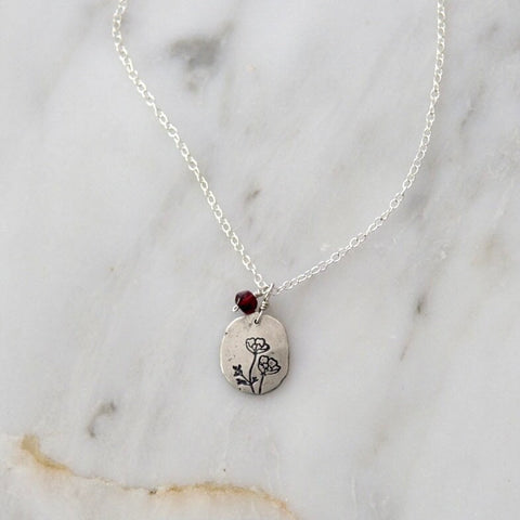 Silver hand stamped poppy pendant with a small attached garnet and a silver chain on a marble backdrop.