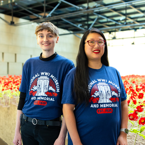 Two people wearing a dark blue t-shirt that depicts the Liberty Memorial Tower with poppy flowers. Arching over the tower is the text 'National WWI Museum and Memorial'. There are poppy flowers in the background.