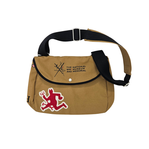 A canvas satchel with a dark strap. on the front is a patch with a red devil insignia patch on the bottom left. On the opening flap is the logo for the National WWI Museum and Memorial.
