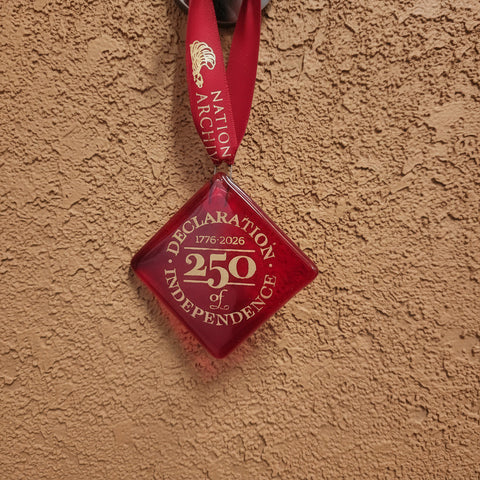 A square ornament made out of red glass. It has gold text that reads, "Declaration of Independence, 250". It has a red ribbon for the National Archives.