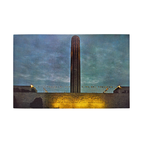 A postcard depicting a photograph of the Liberty Memorial Tower at Dusk. There are flags around the base, with lights shining of the frieze below it.