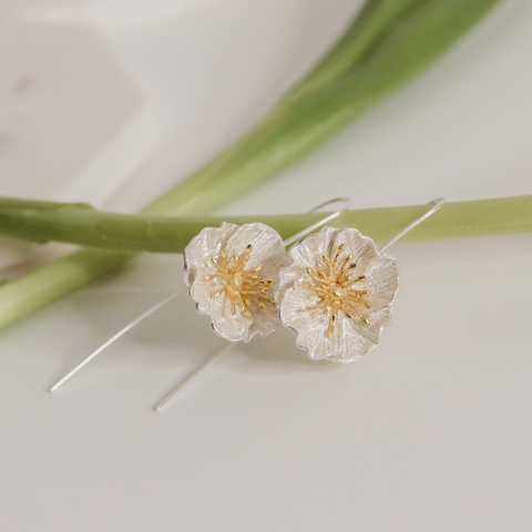 Drop earrings laying on the stem of a flower. They are silver and gold colored and depict poppy flowers.