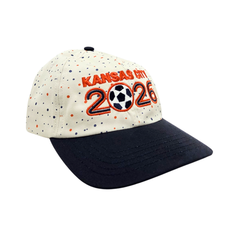 A cream colored baseball hat with a black brim/ On the front is Orange text saying Kansas City 2026, with the 0 in the shape of a soccer ball. The hat has small dark blue and orange polka dots all over it.