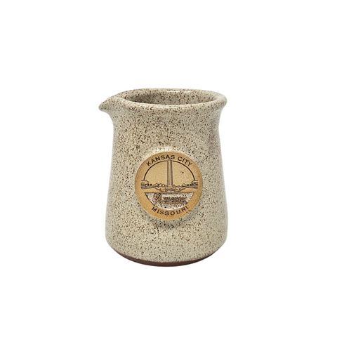 A small pitcher to hold cream and other liquids. It is tan sandy color with speckles of brown on the glaze. In the middle in an emblem which depicts the WWI Musem and text that reads Kansas City Missouri.