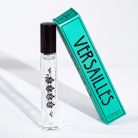 A photo of a 10 ml glass perfume bottle with black floral motifs on the side.  It has a black cap and is standing upright. the box it comes in is leaning against it.