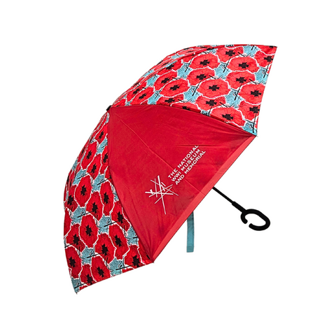 An Image of an opened umbrella with a front panel that has the National WWI Museum and Memorial logo on it. The front panel is red, and the rest of the umbrella is mostly red with digitally manipulated poppies that are repeated and stacked on one another with the National WWI Museum and Memorial Intersections logo interlaced between. the background of these poppies are a cool blue color. The handle is black and has a unique c shaped end.