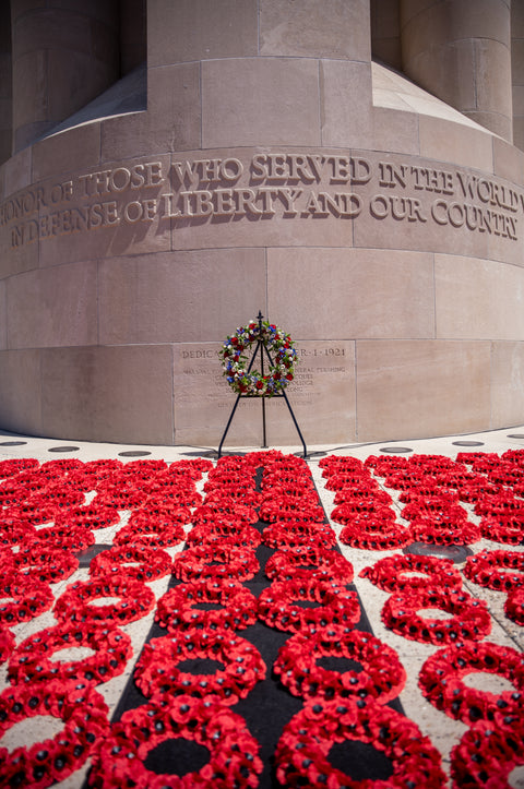 Wreath on a stand in front of a memorial with red poppy wreaths on the ground.