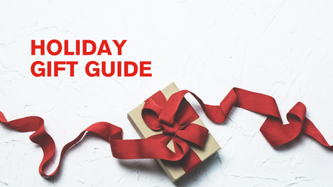 A white banner that has the text Holiday Gift Guide in all caps and in red. The text is in the top right. There is a red ribbon laid out in a decorative manner that spreads across the bottom of the banner, with a wrapped present in the middle.